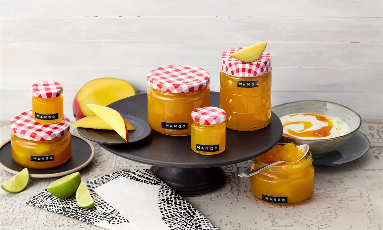 Kozarčki z živahno oranžno mango marmelado, pokriti z belimi in rdečimi karirastimi pokrovčki, na črnem podstavku z rezinami manga