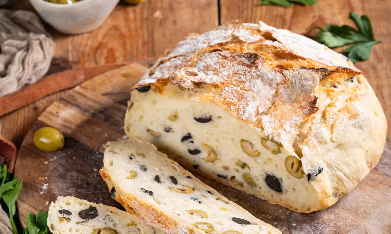 Svetli kruh z zlato rjavo skorjo, razpršeno črnimi in zelenimi olivami, postrežen na leseni deski z vejicami peteršilja