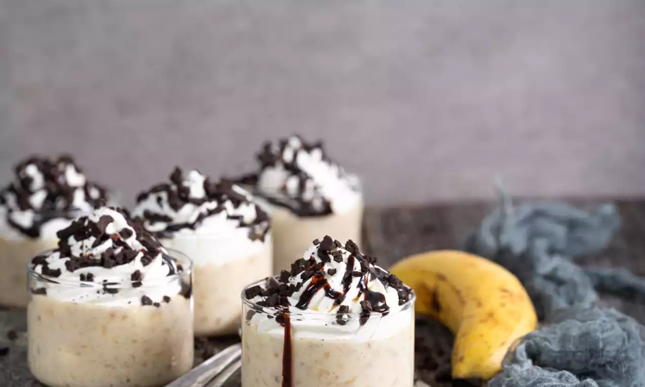 Kremast bananin puding v prozorni skodelici, obložen s stepeno smetano, čokoladnimi koščki in prelit s čokoladnim prelivom na rustikalni deski