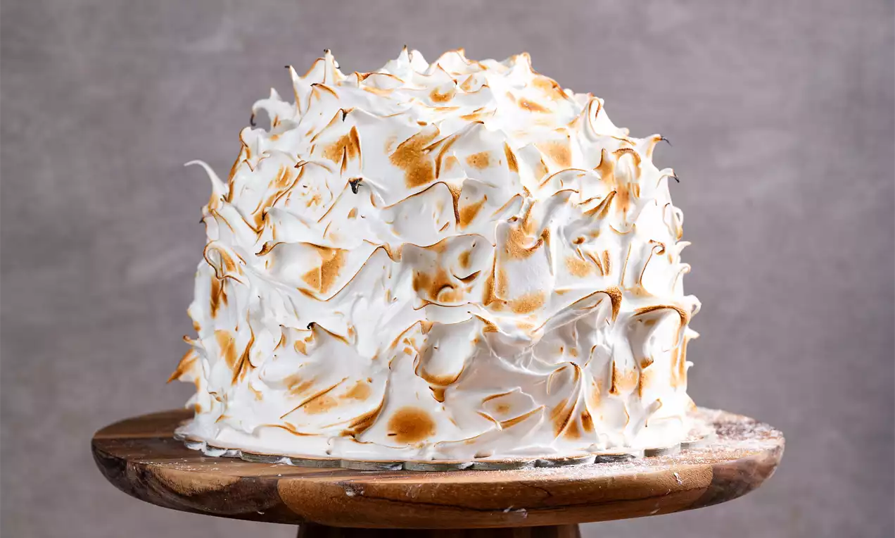 Kupolasta sladica s puhasto belim meringue prekritjem, zlatorjavo karamelizirana na robovih, na leseni podstavku, obdana z borovnicami in dekorjem