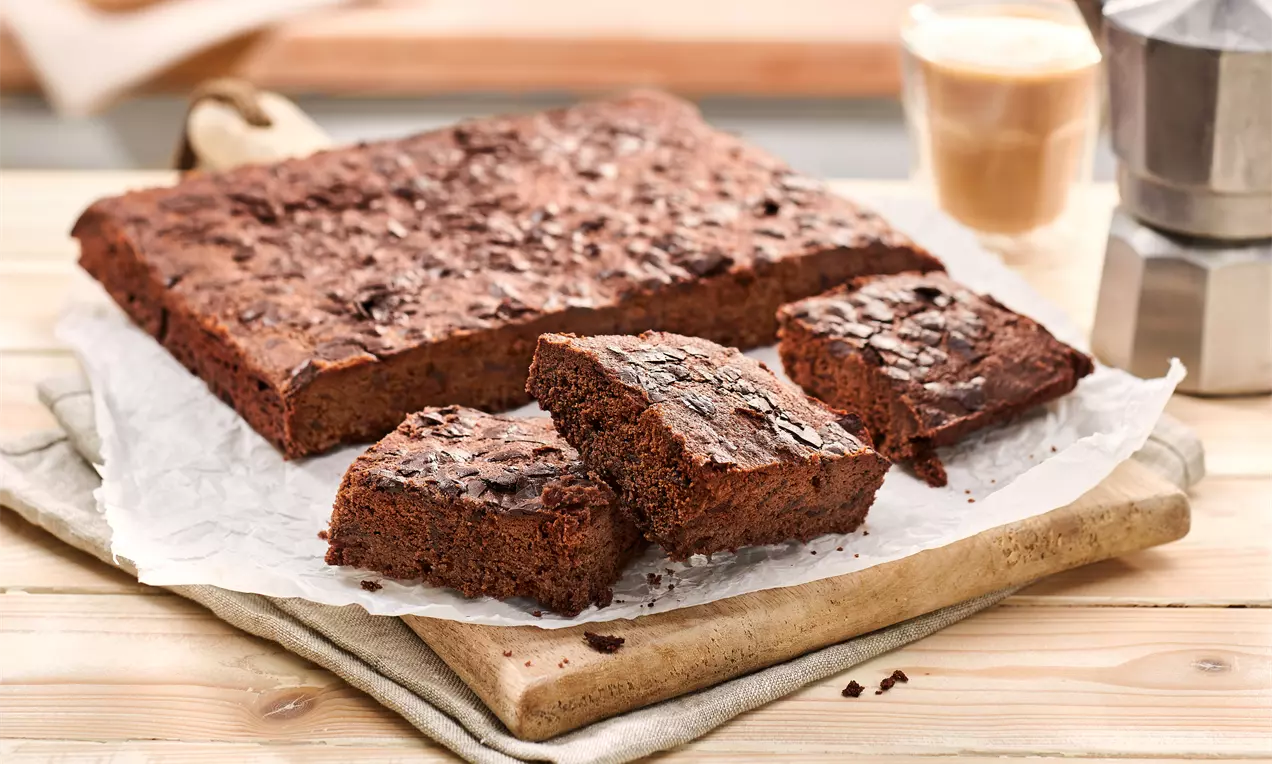 Četvrtasti smeđi brownies s teksturom pukotina na površini, posluženi na papiru za pečenje na drvenoj dasci s čašom kafe u pozadini