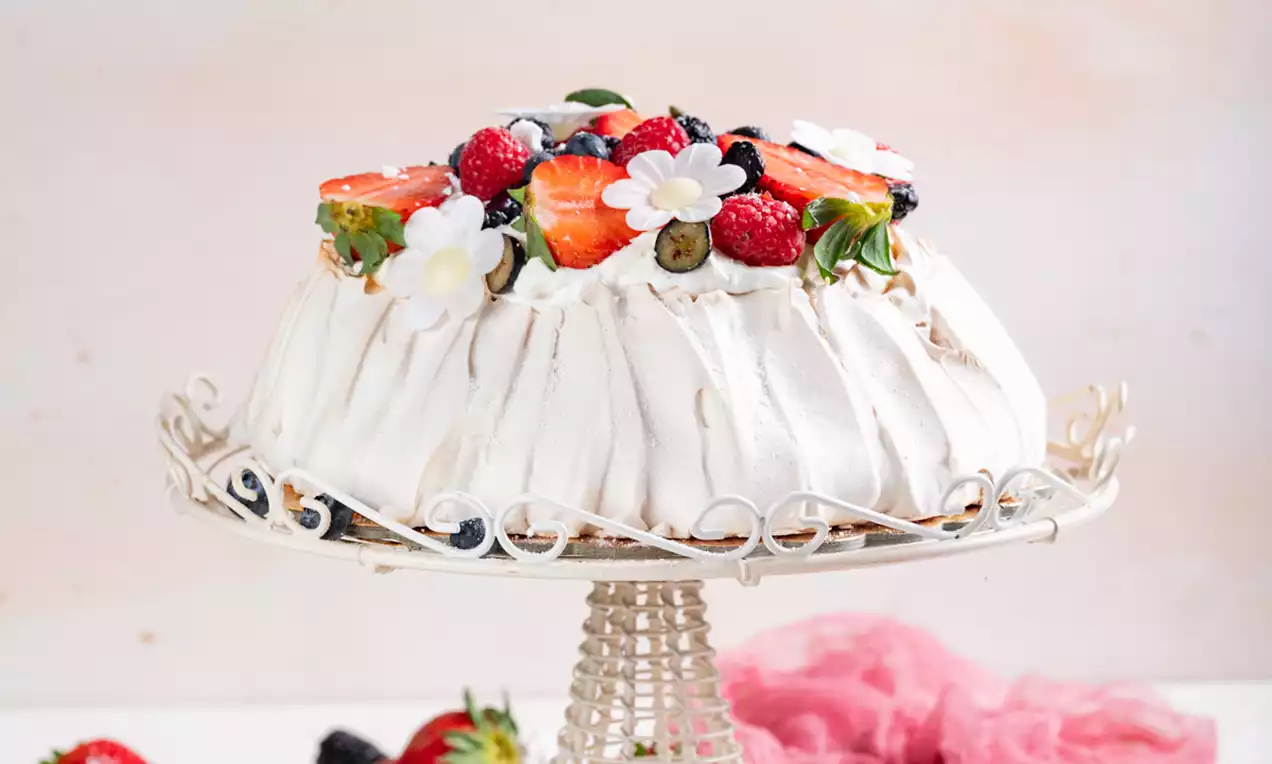 Pavlova snežno bele barve z rahlo nagubano teksturo, okrašena z rdečimi jagodami, borovnicami, cvetovi in pistacijami na elegantnem podstavku