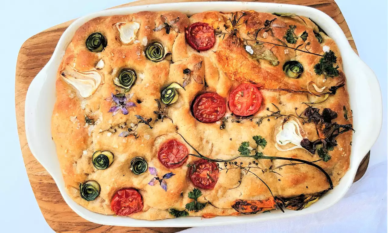 Zlatorjava poletna focaccia z zapečenimi robovi, obložena s pisanimi paradižniki, bučnimi spiralami, zelišči in drobnimi cvetovi na beli posodi