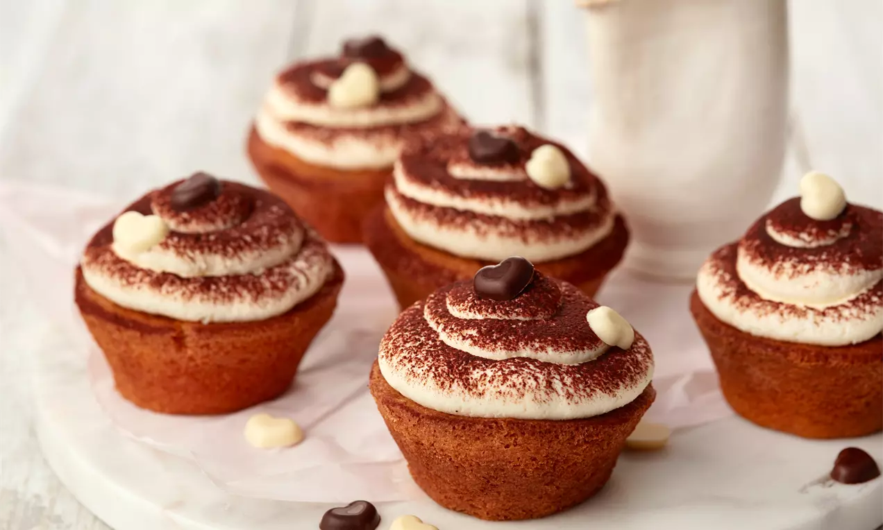 Tiramisu cupcakes z zlato rjavo bazo, svetlo kremo s kakavovim posipom ter čokoladnimi srci, razporejeni na belem krožniku