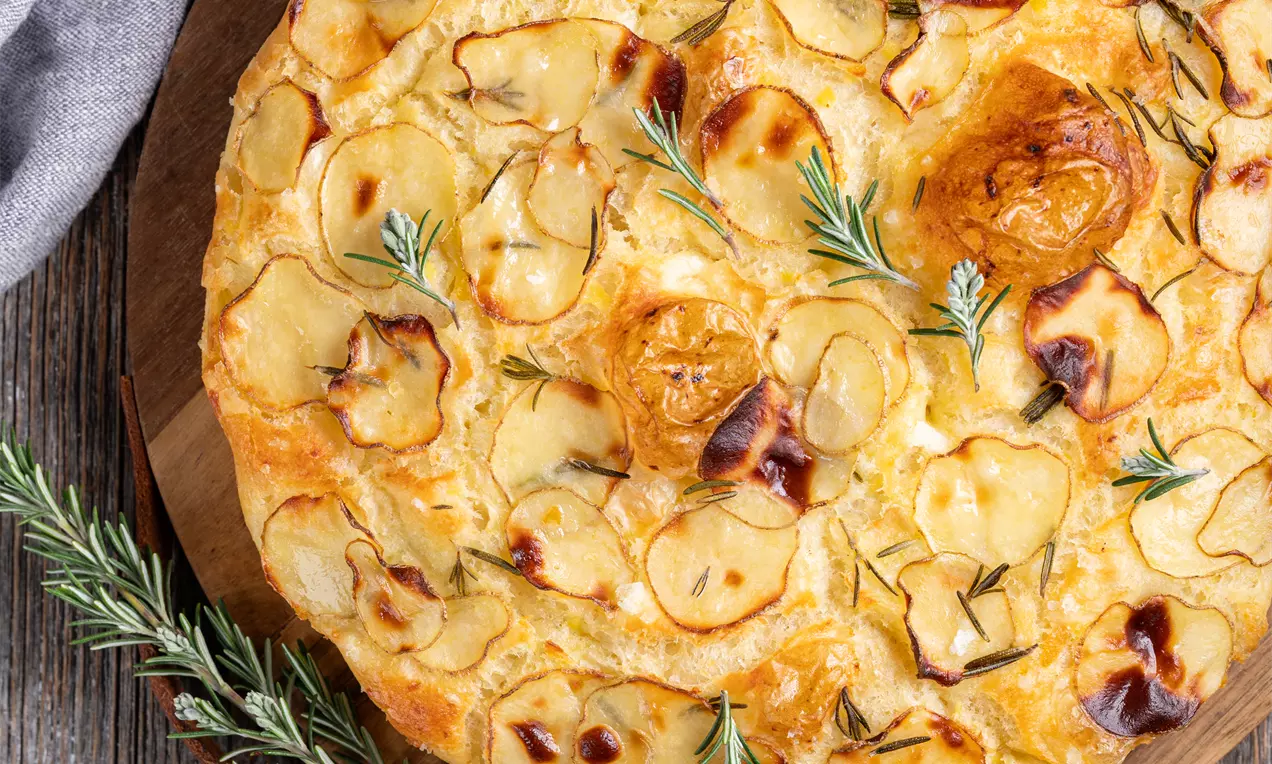 Zlato-rjava krompirjeva focaccia z rezinami krompirja, hrustljavo skorjo, vejicami rožmarina na leseni deski in rustikalnem ozadju