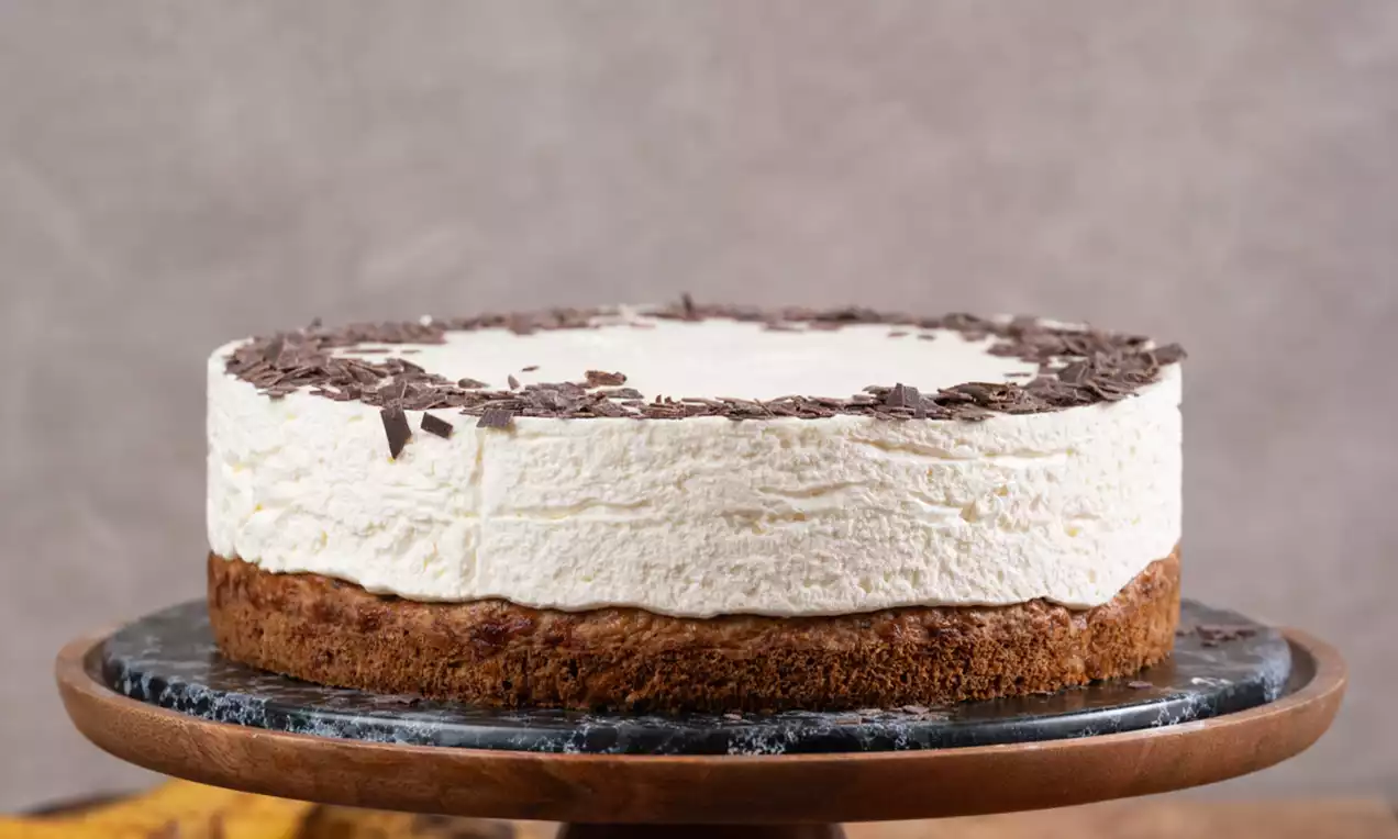 Cheesecake z rahlo kremasto plastjo svežega sira na vlažni bazi iz bananinega kruha, posut z drobnimi čokoladnimi ostružki na rjavem podstavku