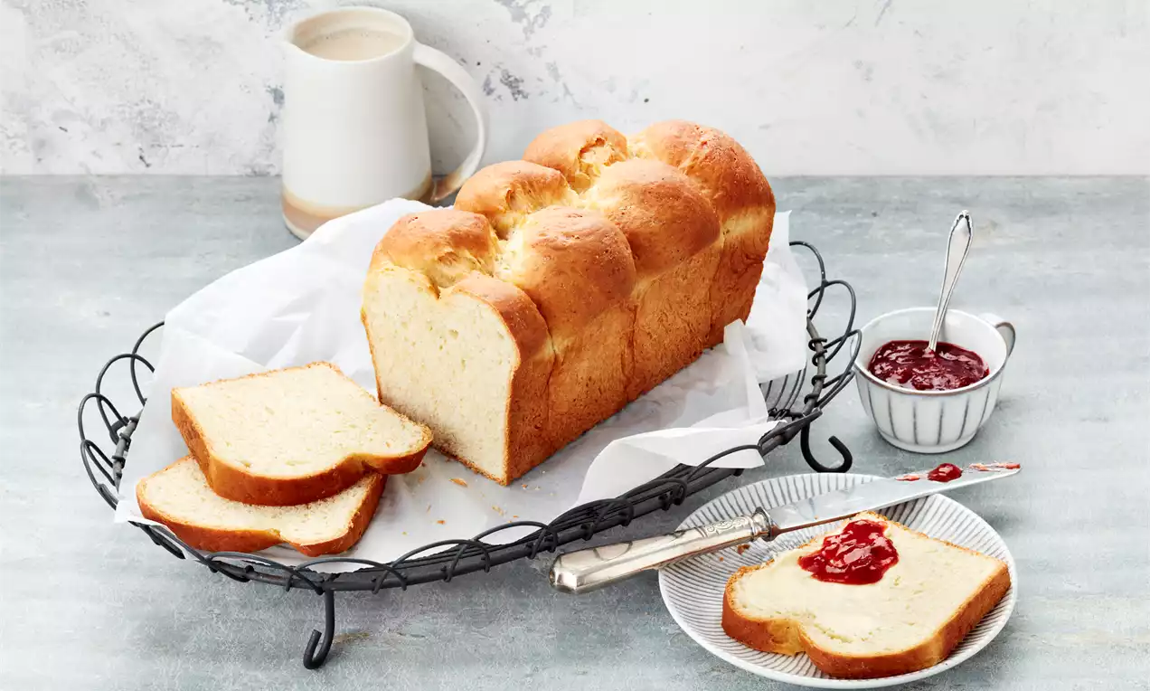 Zlatorumena brioš z mehko in puhasto teksturo, razrezana na enakomerne rezine, postrežena na črnem pladnju z marmelado in skodelico čaja