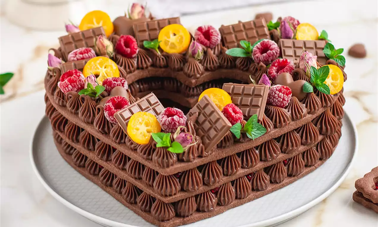 Srčasta čokoladna torta iz temno rjavih plasti, okrašena s čokoladnimi kremnimi venci, svežimi malinami, rezinami kumkvata, metinimi listi in čokoladnimi kvadratki