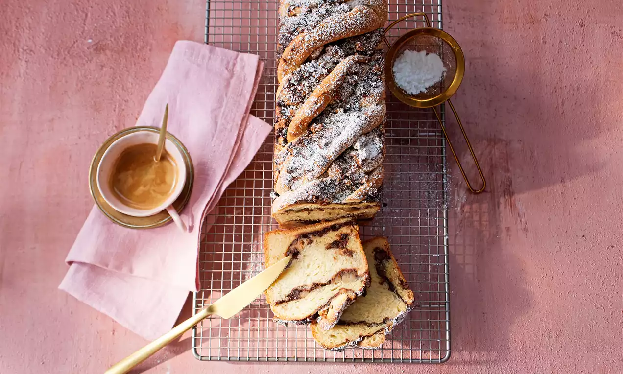 Babka s plastmi rahlega testa in temnega nadeva, spiralno zavita, posuta s sladkorjem v prahu na mreži ob roza servetu in skodelici kreme