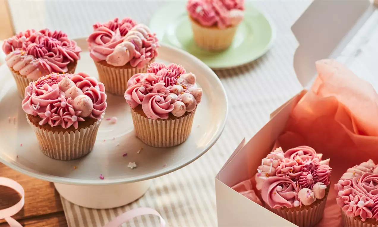 Rožnati cupcake-i s skrbno nabrizgano kremo v vzorcih rož, okrašeni s perlami in drobnimi sladkimi dodatki na belem pladnju