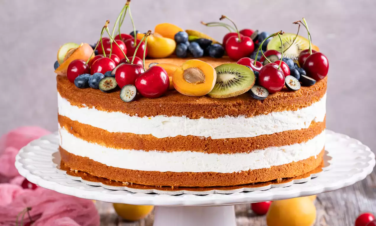 Skutina torta z zlatimi biskvitnimi plastmi in snežno belo kremo, obložena z jagodami, češnjami, marelicami in kivijem na belem podstavku