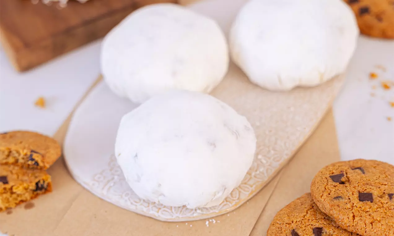Beli puhasti mochi piškoti z gladko površino, položeni na svetleč pladenj, obkroženi s hrustljavimi rjavimi piškoti in drobtinami.