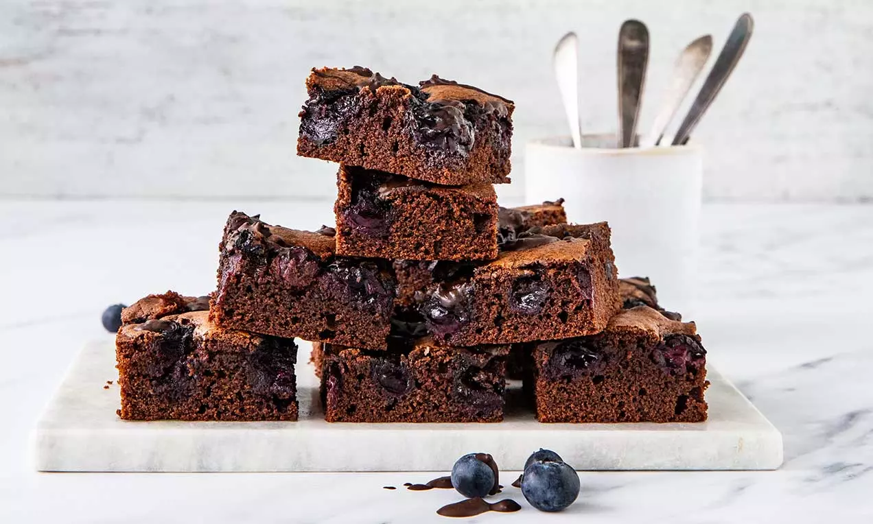 Četvrtasti brownies tamnosmeđe boje prošarani sočnim borovnicama, složeni u piramidu na bijeloj plohi s nekoliko svježih borovnica