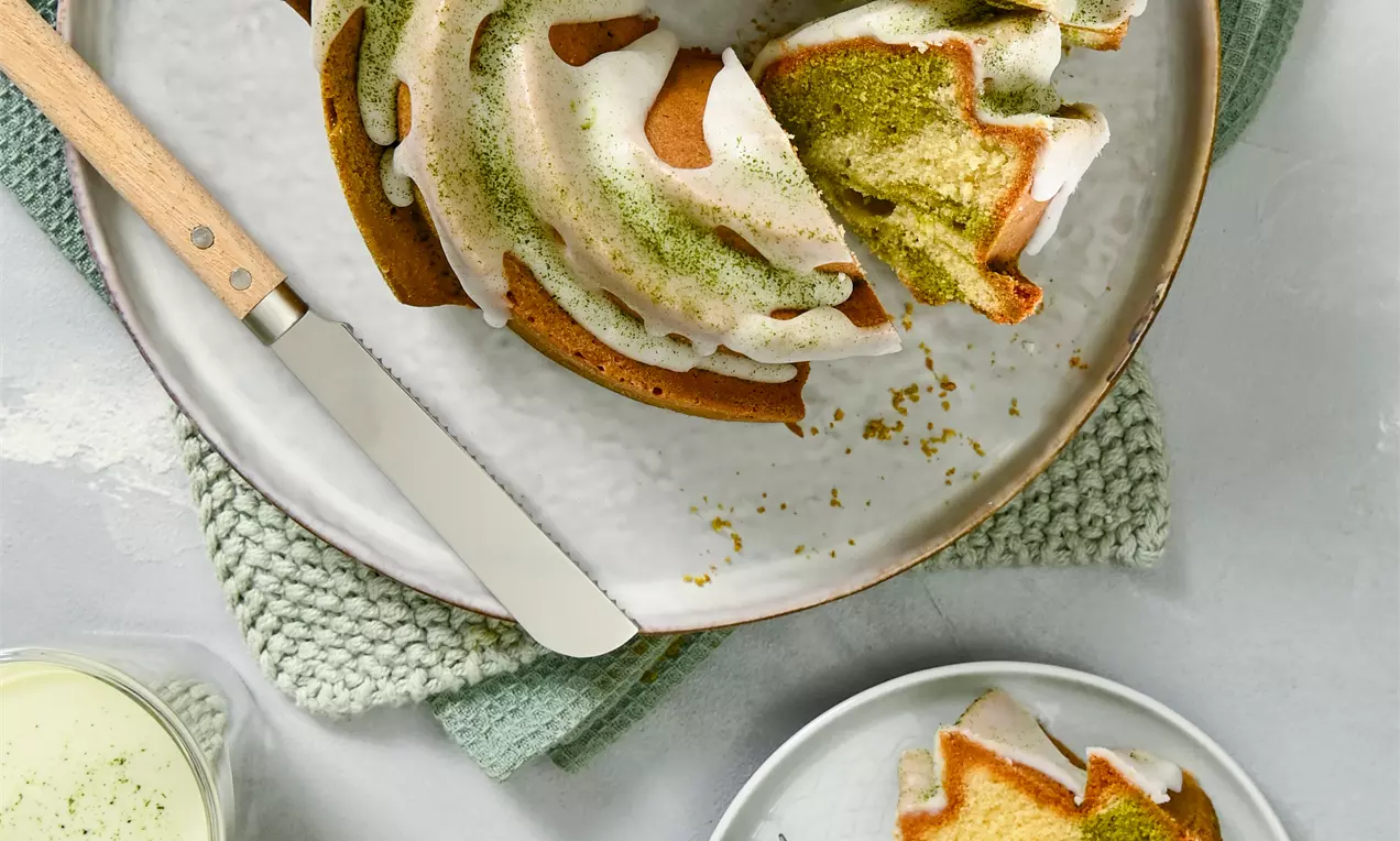 Bundt kolač s spiralno obliko, dvobarvnim notranjim vzorcem zelenega matcha prahu in vanilijevega testa, prelit z glazuro in potresen z matcha prahom